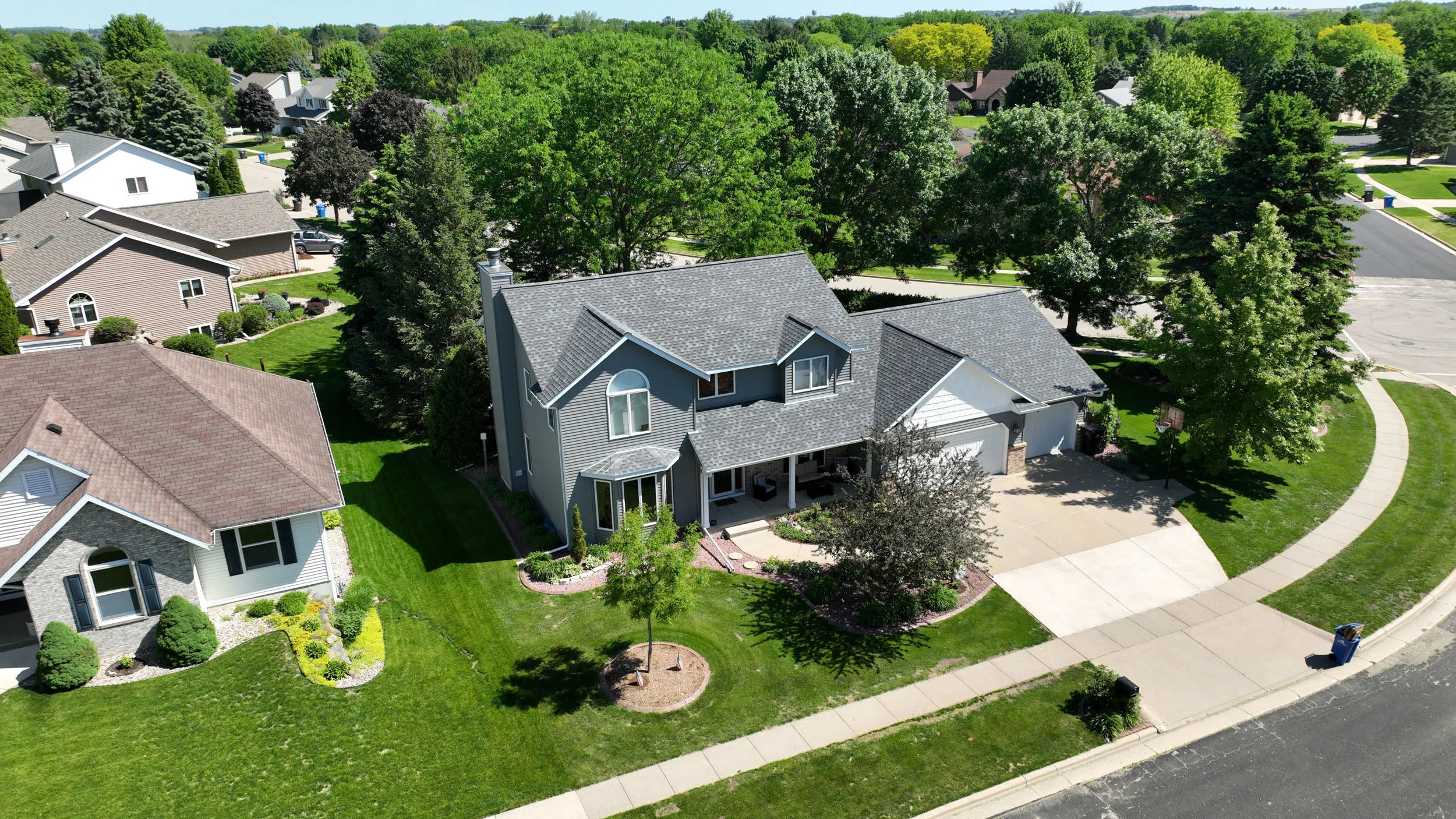 Aerial view of a two-story gray house with a well-maintained lawn, surrounded by trees and neighboring houses on a sunny day.